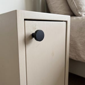 Pu&ograve; includere: Un comodino in legno color beige chiaro dotato di un'anta singola con un pomello circolare nero opaco e liscio. Il mobile presenta una struttura rettangolare e una venatura del legno visibile, offrendo una soluzione di archiviazione.