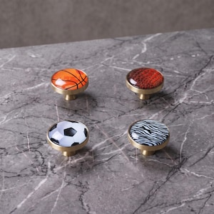 Könnte beinhalten: Vier runde Schrankknöpfe mit einem goldfarbenen Metallsockel und einer gewölbten Oberseite. Die Designs umfassen einen orangefarbenen Basketball, ein braunes Strukturmuster, einen schwarz-weißen Fußball und ein schwarz-weißes Zebramuster.