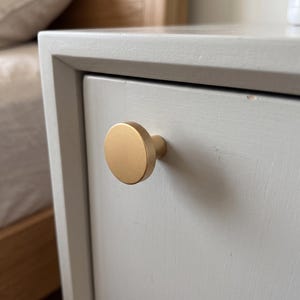 Pu&ograve; includere: Un pomello per mobili circolare in oro opaco con una faccia piatta e una base cilindrica, montato su un frontale di cassetto in legno verniciato grigio chiaro. L'hardware presenta una finitura metallica liscia e un design geometrico.