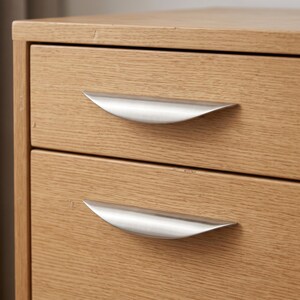 May include: A light oak wood chest of drawers featuring two stacked drawers. Each drawer is equipped with a curved, brushed silver metal handle that spans the centre width of the drawer front, showcasing a natural wood grain texture throughout.