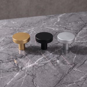 Pode incluir: Três puxadores circulares para armários com um acabamento liso e mate. O conjunto inclui um puxador em tom dourado, um preto mate e um em tom prateado. Cada puxador apresenta uma superfície superior redonda e plana com uma base cilíndrica.
