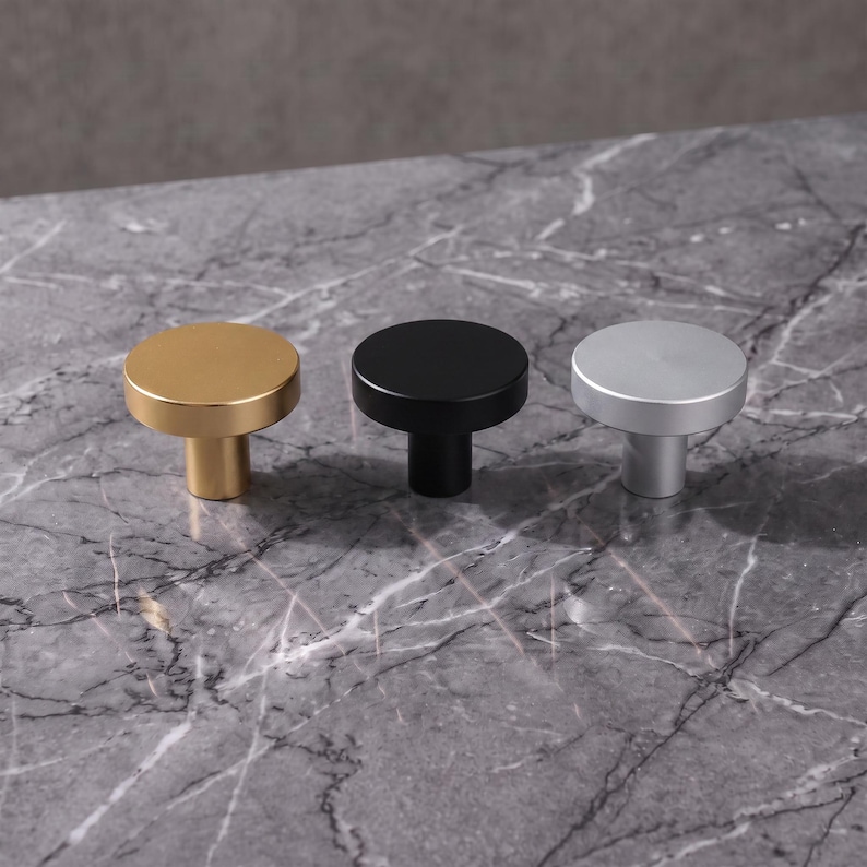 Pu&ograve; includere: Tre pomelli circolari per mobili con una finitura liscia e opaca. Il set include un pomello color oro, uno nero opaco e uno color argento. Ogni pomello presenta una superficie superiore rotonda e piatta con una base cilindrica.