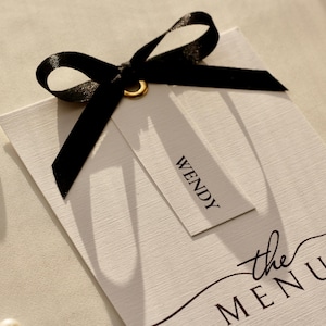 Könnte beinhalten: Eine weiße Menükarte mit dem Text "the MENU" und dem Namen "WENDY" auf einem Anhänger, verziert mit einem schwarzen Band. Die Karte hat eine strukturierte Oberfläche und ist mit einem goldenen Ring akzentuiert. Zwei Perlen sind sichtbar.