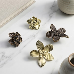 Op de afbeelding: Vier decoratieve bloemvormige kastknoppen in goud- en bronskleuren. De metalen knoppen hebben gedetailleerde bloemblaadjes. Ze zijn gerangschikt op een wit marmeren oppervlak, wat wijst op een interieurthema.