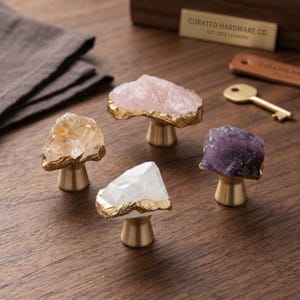 May include: Four decorative cabinet knobs with natural crystal tops in shades of pink, yellow, white, and purple. Each knob has a gold-colored metal base. The knobs are arranged on a wooden surface.