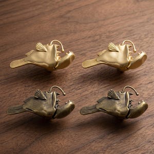 Könnte beinhalten: Vier Schrankknöpfe in Form von Anglerfischen aus Messing und Bronze. Die Knöpfe haben offene Münder, scharfe Zähne und einen leuchtenden Köder. Zwei sind goldfarben und zwei bronzefarben, alle mit einem Used-Look. Die Knöpfe sind auf einer Holzoberfläche angeordnet.