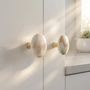 Può includere: Due pomelli ovali per armadi con un design marmorizzato nei toni crema, pesca e oro. Le manopole sono montate su hardware in ottone e sono attaccate a un'anta bianca. Le manopole sono un accento decorativo.