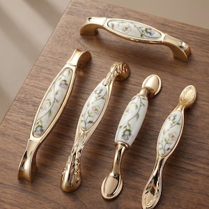 May include: A collection of gold-toned cabinet pulls and handles. Each handle features a white ceramic center with a delicate floral pattern in pastel colors. The hardware is designed with ornate details and a polished finish.