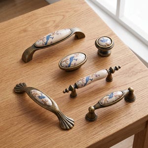 Può includere: Una collezione di ferramenta per mobili in stile antico, tra cui maniglie e un pomello. La ferramenta presenta un centro in ceramica color crema con un motivo floreale e uccelli blu, incastonato in una cornice di metallo color bronzo. Le maniglie variano per dimensioni e forma.