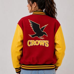 Könnte beinhalten: Eine Collegejacke mit einem dunkelroten Wollmischkörper und leuchtend gelben Ärmeln. Auf der Rückseite befindet sich eine große schwarze Rabengrafik über dem gelben Schriftzug "CROWS" mit weißer Umrandung. Sie hat gerippte Kragen und Ärmelbündchen.