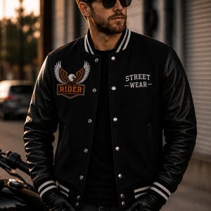May include: Black varsity jacket with leather sleeves, white stripes on the cuffs and collar, and the text "STREET -WEAR-". An embroidered eagle patch and the word "RIDER" are on the chest. The jacket is worn with black gloves and sunglasses.
