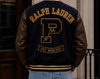 Könnte beinhalten: Eine marineblaue Collegejacke mit braunen Lederärmeln und gestreiften Rippbündchen. Auf der Rückseite befindet sich eine gelbe Stickerei mit RALPH LAUREN, einem großen Buchstaben P mit dem Wort Polo und einem Banner mit EST. MCMLXVII.
