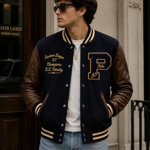 Puede incluir: Una chaqueta universitaria azul marino con mangas de cuero marrón, con ribetes acanalados a rayas color crema en el cuello, puños y dobladillo. El frente muestra un bordado dorado que dice Eastern League '67 Champions R.L. Varsity y una letra P grande.