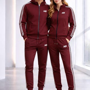 Puede incluir: Conjuntos de chándal burdeos a juego con rayas blancas y las letras "NS" en el pecho y la pierna. Los chándales incluyen una chaqueta con capucha y cremallera y pantalones a juego. Los modelos llevan zapatillas blancas.