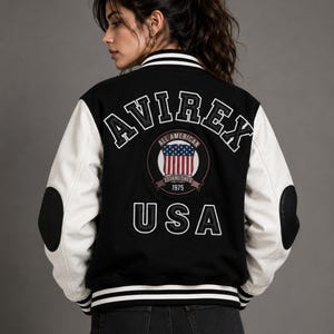 Female model wearing black and white varsity jacket back view with oversized embroidered AVIREX logo