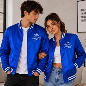 Matching couple wearing royal blue satin varsity bomber jackets in modern indoor streetwear lifestyle setting.
