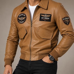 Puede incluir: Una chaqueta de cuero color canela con cremallera frontal, puños acanalados y dos bolsillos con solapa. Incluye tres parches: uno circular 'VINTAGE AIR DIVISION', uno rectangular 'SKY RIDERS FLIGHT CLUB' y uno en forma de escudo 'AIR COMMAND 79'.