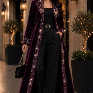 May include: A long, deep purple velvet coat featuring wide lapels and cuffs heavily embellished with sparkling crystals. The coat is worn over a black top and black trousers with a belt, paired with a small black handbag and dark sunglasses.