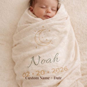 Op de afbeelding: Een gepersonaliseerde babydeken in crèmekleur, gewikkeld om een slapende baby. De deken heeft een maan- en sterrenontwerp, de naam "Noah" en de datum "02.20.2026". De tekst "Custom Name + Date" is ook zichtbaar.