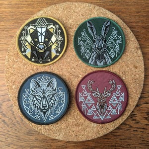 Peut inclure: Quatre écussons ronds brodés avec des motifs géométriques d'animaux. Les écussons représentent un blaireau, un lapin, un loup et un cerf, chacun sur un fond de couleur différente. Les écussons sont présentés sur une surface en liège.