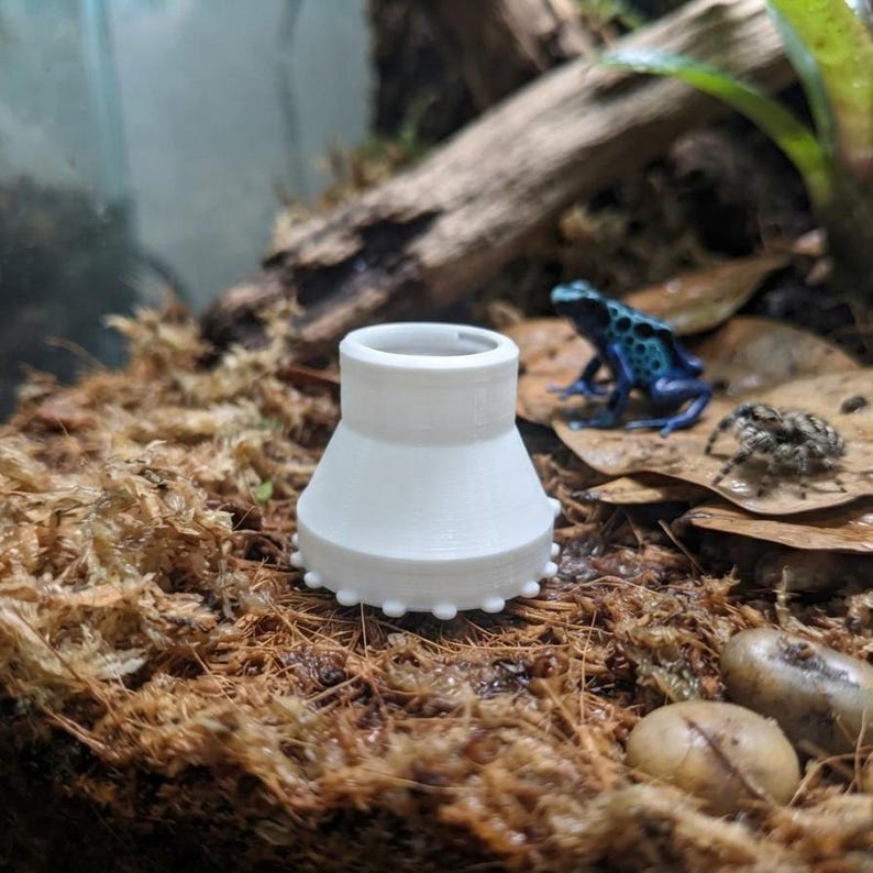 Peut inclure: Un adaptateur conique en plastique blanc imprim&eacute; en 3D, dot&eacute; d'un centre creux et d'un bord de base orn&eacute; de petites protub&eacute;rances circulaires r&eacute;guli&egrave;rement espac&eacute;es. L'objet est plac&eacute; verticalement sur un substrat organique brun.