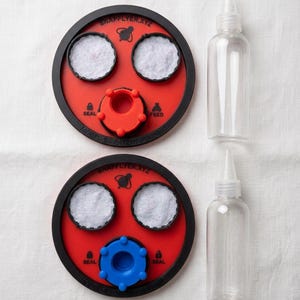 Peut inclure: Deux dispositifs circulaires rouges et noirs marqu&eacute;s SNAPFLYER.XYZ avec des inserts circulaires blancs, l'un avec une pi&egrave;ce centrale rouge et l'autre bleue, accompagn&eacute;s de deux flacons souples transparents &agrave; embout pointu.