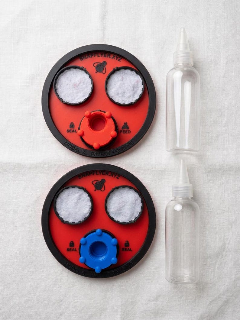 Peut inclure: Deux dispositifs circulaires rouges et noirs marqu&eacute;s SNAPFLYER.XYZ avec des inserts circulaires blancs, l'un avec une pi&egrave;ce centrale rouge et l'autre bleue, accompagn&eacute;s de deux flacons souples transparents &agrave; embout pointu.