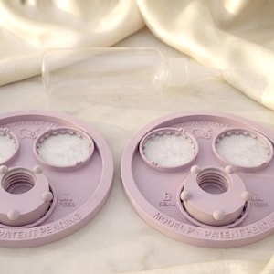 Peut inclure: Deux couvercles circulaires en plastique violet clair avec des inserts blancs textur&eacute;s, chacun dot&eacute; d'une ouverture centrale filet&eacute;e, de petits boutons circulaires et du texte en relief "MODEL P - PATENT PENDING," "SEAL," et "FEED." Une bouteille en plastique transparent est plac&eacute;e derri&egrave;re.