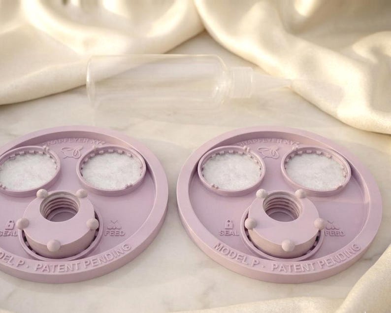 Peut inclure: Deux couvercles circulaires en plastique violet clair avec des inserts blancs textur&eacute;s, chacun dot&eacute; d'une ouverture centrale filet&eacute;e, de petits boutons circulaires et du texte en relief "MODEL P - PATENT PENDING," "SEAL," et "FEED." Une bouteille en plastique transparent est plac&eacute;e derri&egrave;re.