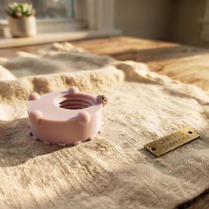 Peut inclure: Un objet rose p&acirc;le, circulaire et filet&eacute; en forme d'&eacute;crou avec de petites protub&eacute;rances arrondies sur le bord ext&eacute;rieur. Une petite araign&eacute;e est pos&eacute;e sur le rebord. &Agrave; c&ocirc;t&eacute; se trouve une &eacute;tiquette m&eacute;tallique rectangulaire grav&eacute;e CRAFTED SNAPFLYER.