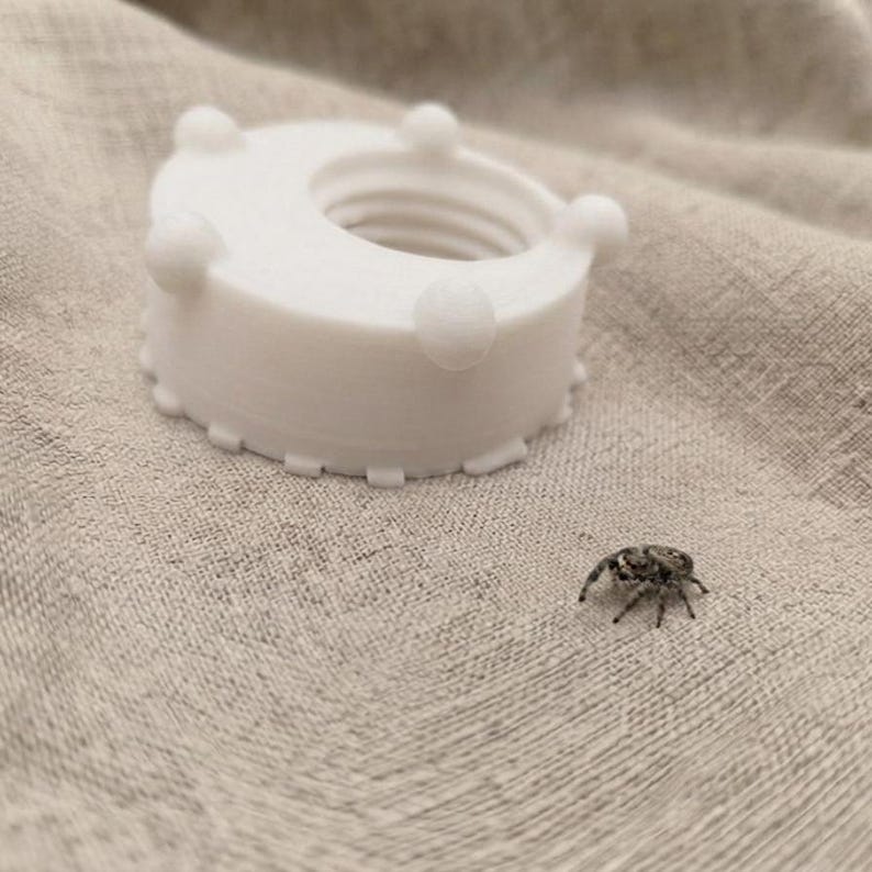 Peut inclure: Un bo&icirc;tier en plastique blanc imprim&eacute; en 3D, de forme circulaire, dot&eacute; d'un trou central filet&eacute; et de quatre petites protub&eacute;rances sph&eacute;riques sur le bord sup&eacute;rieur, plac&eacute; &agrave; c&ocirc;t&eacute; d'une petite araign&eacute;e sauteuse grise sur un tissu beige.