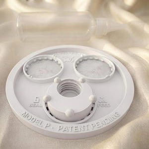 Peut inclure: Un composant circulaire en plastique blanc avec deux zones encastr&eacute;es circulaires, une ouverture centrale filet&eacute;e et le texte en relief SEAL, FEED, et MODEL P. PATENT PENDING. Un flacon souple en plastique transparent est plac&eacute; derri&egrave;re.