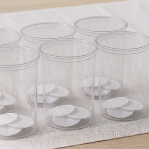 Peut inclure: Six récipients en plastique transparent avec couvercles, chacun contenant plusieurs tampons de coton ronds blancs. Les récipients sont cylindriques et reposent sur une surface blanche texturée. L'image est bien éclairée et montre les produits de manière propre et organisée.