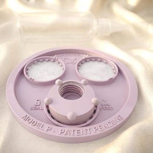 Peut inclure: Un couvercle circulaire en plastique violet clair avec une ouverture centrale filet&eacute;e, deux inserts de filtre circulaires et un texte en relief indiquant SEAL, FEED, et MODEL P. PATENT PENDING. Un flacon souple transparent est plac&eacute; derri&egrave;re.