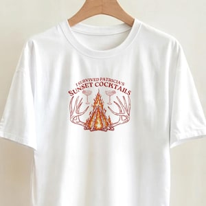 May include: A white short-sleeve t-shirt featuring a central graphic of a campfire flanked by two deer antlers and two cocktail glasses. The text above the fire reads I SURVIVED PATRICIA'S SUNSET COCKTAILS in a red, arched, serif font.