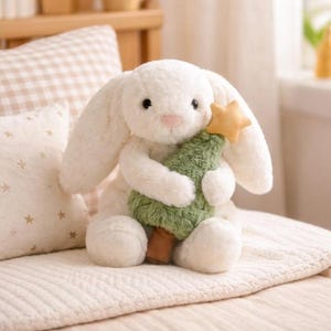 May include: A plush white bunny holding a green Christmas tree with a gold star on top. The bunny has long ears, a pink nose, and black eyes. The tree has a brown trunk. The bunny is sitting on a cream-colored blanket.