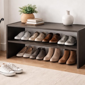 May include: A dark brown shoe rack with two shelves, displaying various shoes. The top shelf holds sneakers and dress shoes in shades of gray, tan, and brown. The bottom shelf features boots and flats. A pair of white sneakers sits on the floor.
