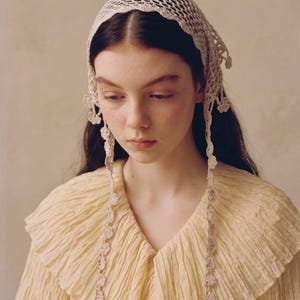 May include: A portrait featuring a person wearing a cream-colored, crocheted head covering and a light yellow, ruffled blouse. The head covering has a scalloped edge and floral accents. The blouse has a textured, pleated appearance.