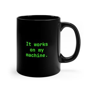 Può includere: Una tazza in ceramica nera con il testo "It works on my machine." stampato in un colore verde lime brillante con un carattere a spaziatura fissa. La tazza presenta una finitura liscia e lucida, un corpo cilindrico e un manico curvo.