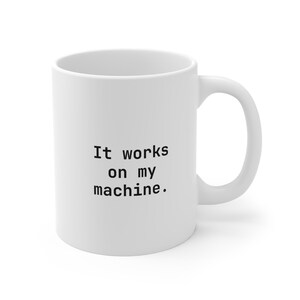 May include: A white ceramic mug featuring the black text It works on my machine. printed in a monospaced font on the front. The mug has a smooth finish, a cylindrical body, and a curved handle on the right side, providing a simple and functional design.