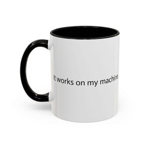 May include: A white ceramic mug with a black interior and a black handle. The exterior features the black printed text "It works on my machine." The mug has a cylindrical shape with a smooth finish and a curved handle on the left side.