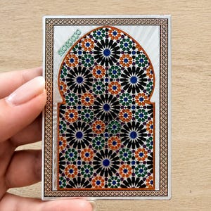 Könnte beinhalten: Eine rechteckige Karte mit einem bogenförmigen Design, das mit einem geometrischen Mosaikmuster in Schwarz, Blau, Grün und Orange gefüllt ist. Das Wort MOROCCO ist oben links in Grün gedruckt. Die Karte hat einen dekorativen Rand mit Muster.