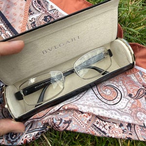 Op de afbeelding: Een bril van Bvlgari met dunne zilverkleurige metalen monturen en zwarte pootjes, rustend in een zwarte harde brillenkoker. De binnenkant van de koker is bekleed met beige stof en is in het midden voorzien van de merknaam Bvlgari.