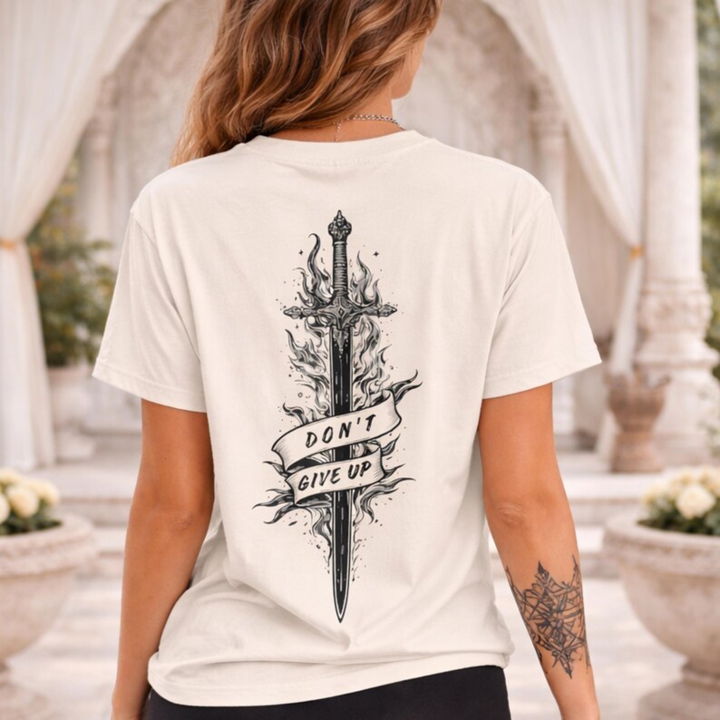 May include: Cream-colored t-shirt featuring a black sword design with flames and a banner that reads "DON'T GIVE UP." The graphic is centered on the back of the shirt. The shirt is made of a soft material.