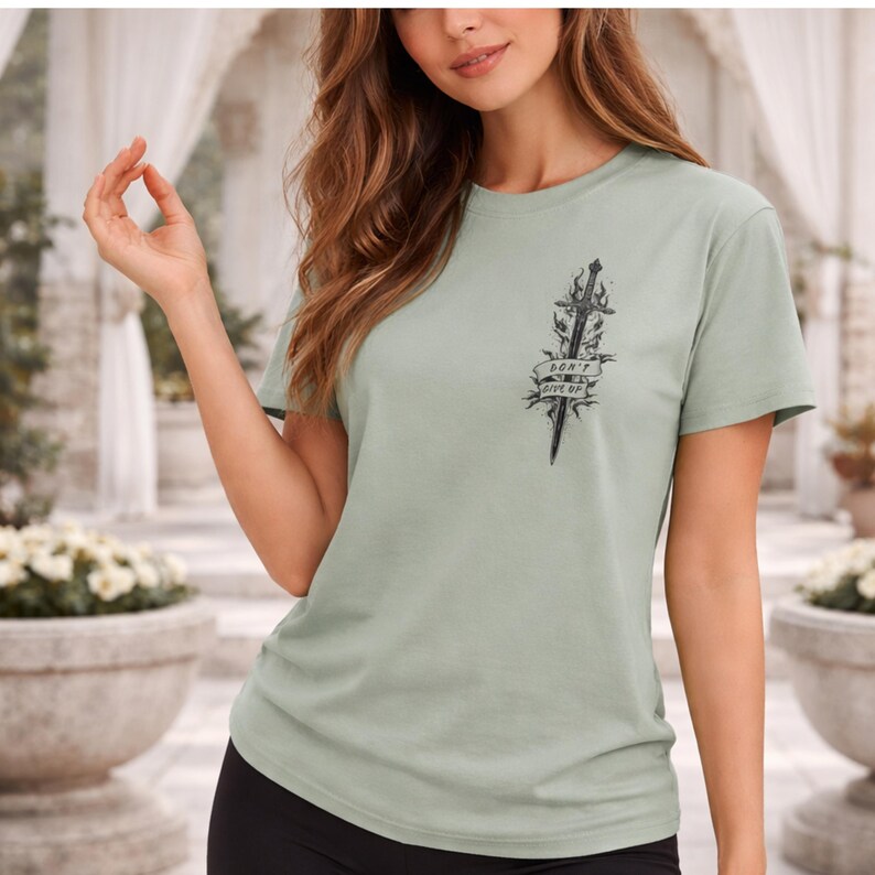 May include: Sage green t-shirt with a black graphic of a sword and the words "DON'T GIVE UP". The short-sleeved shirt is made of a soft material and has a classic crew neck design. The graphic is centered on the chest.