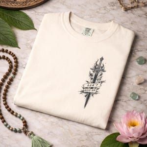 May include: Cream-colored t-shirt featuring a black and gray sword design with flames and the text "DON'T GIVE UP" on a banner. The shirt is folded and displayed on a marble surface.