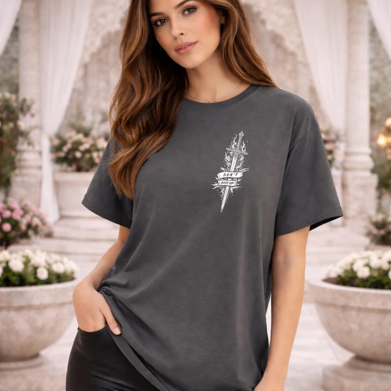 May include: Dark gray t-shirt with a white graphic of a sword and flames, and the text "DON'T GIVE UP". The short-sleeved shirt is made of a soft material.