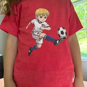 Puede incluir: Camiseta roja con una ilustraci&oacute;n de dibujos animados de un jugador de f&uacute;tbol pateando un bal&oacute;n de f&uacute;tbol. El jugador lleva un uniforme blanco y azul. La camiseta est&aacute; hecha de un material suave y c&oacute;modo.
