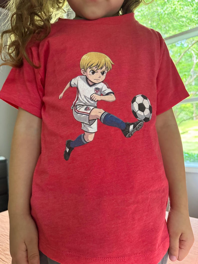 Puede incluir: Camiseta roja con una ilustraci&oacute;n de dibujos animados de un jugador de f&uacute;tbol pateando un bal&oacute;n de f&uacute;tbol. El jugador lleva un uniforme blanco y azul. La camiseta est&aacute; hecha de un material suave y c&oacute;modo.