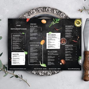 May include: A black chalkboard-style restaurant menu with white text, featuring sections for noshes, soups, fresh salads, specials, boards, herring, sweets, and platters. The menu is on a silver platter with a fork and knife.